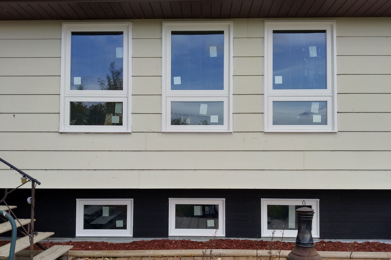 Newly installed white-framed windows on a beige house, with three large double-hung windows on the upper level and three smaller windows on the lower level, surrounded by a freshly landscaped garden bed.